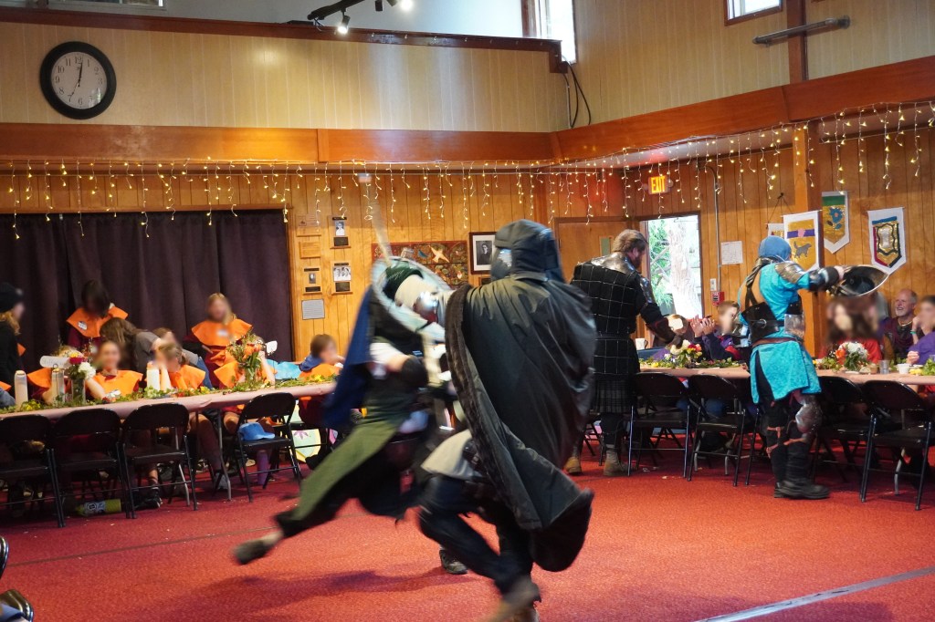 Cut-and-thrust sword fighters in motion, while heavy fighters show off their gear to kids in the background at Siskiyou School in Ashland. April 16, 2026. Kids' faces are blurred for identity protection.