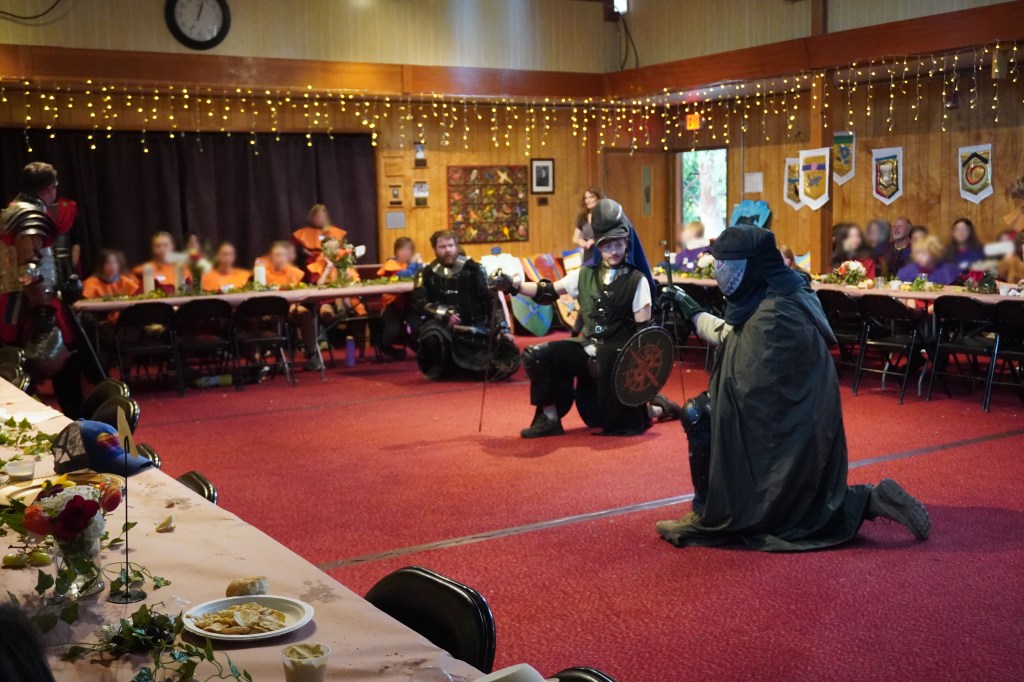 John Latson, Cody Williams and Conner Hummel kneel with respect while the king and queen address the room at Siskiyou School in Ashland, Oregon. April 16, 2026. Kids' faces are blurred for identity protection.